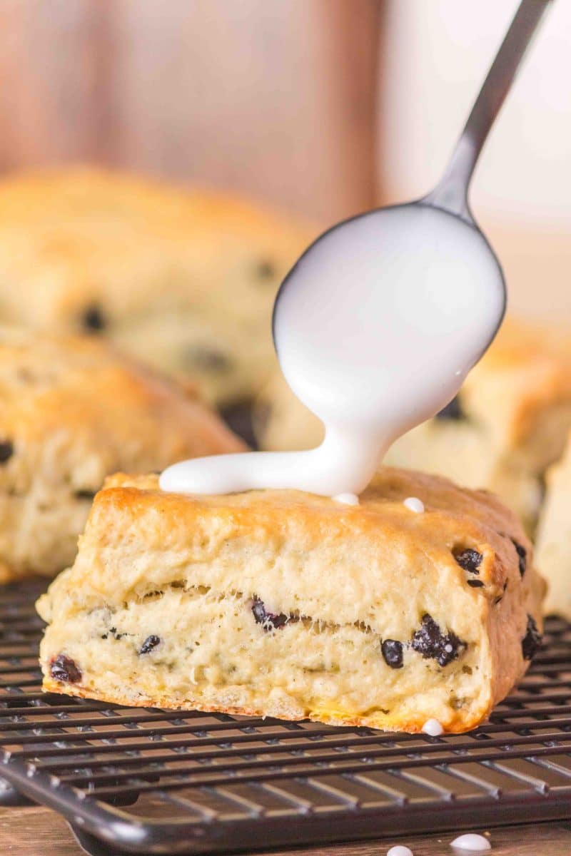 Vanilla glaze being drizzled over a blueberry biscuit sitting on a cooling rack. Hostess At Heart