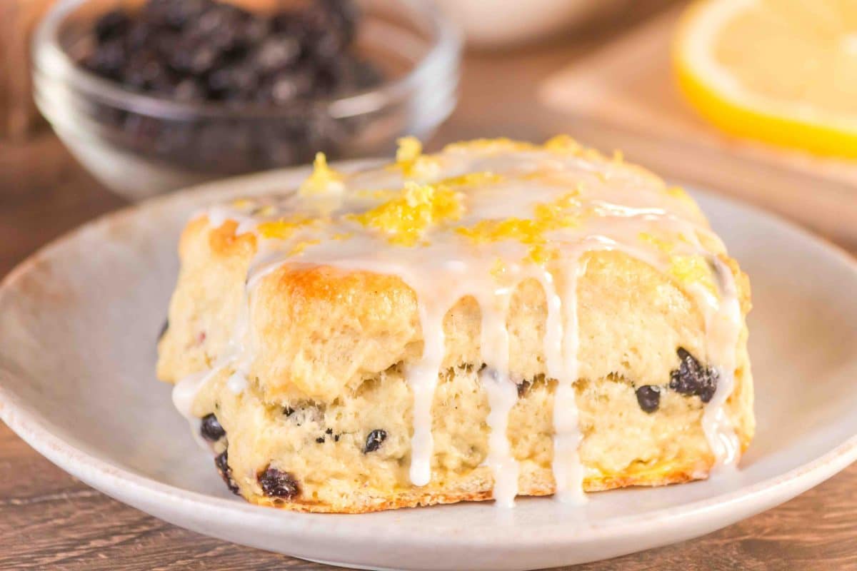 Angled view of a homemade blueberry biscuit topped with a vanilla glaze drizzle and zested lemon on a plate. Hostess At Heart