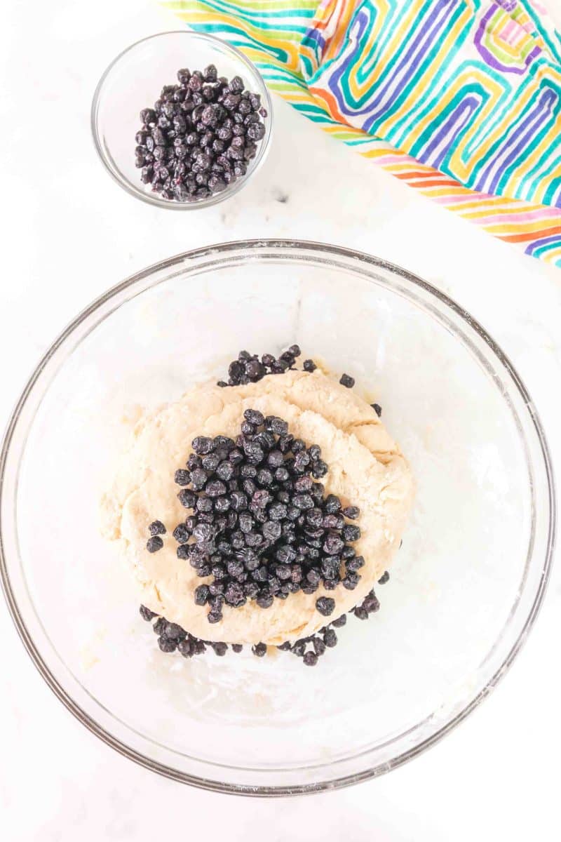 Overhead view of a mixing bowl containing blueberry biscuit recipe dough with topped with dried blueberries ready to be folded. Hostess At Heart
