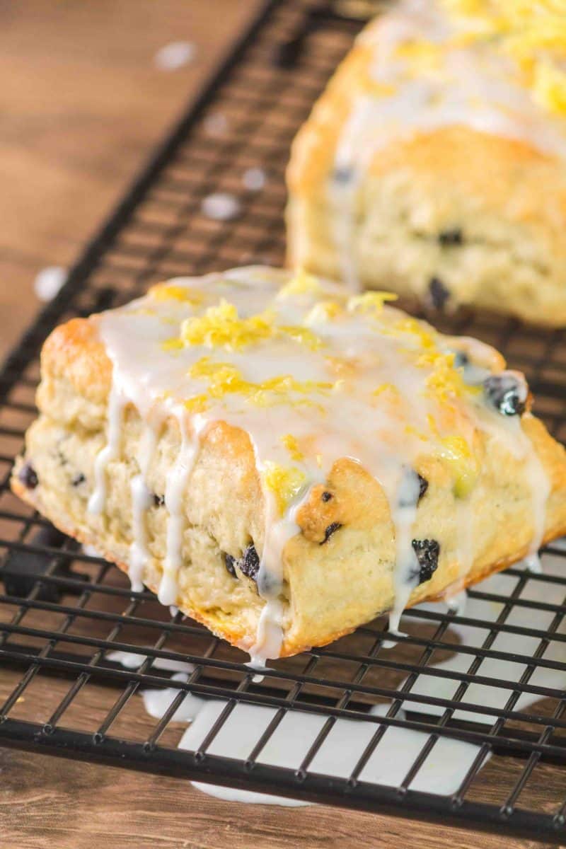 Sideview of a freshly glazed blueberry biscuit on a cutting rack garnished with lemon zest. Hostess At Heart