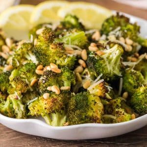 Golden oven roasted broccoli garnished with browned pine nuts and Parmesan cheese in a large serving dish. Hostess At Heart