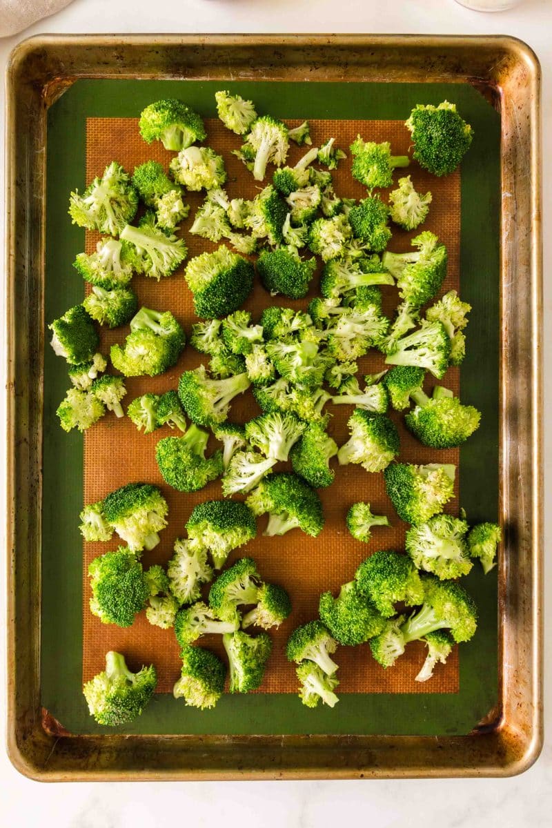 Seasoned broccoli spread in a single layer on a lined baking sheet ready to roast. Hostess At Heart