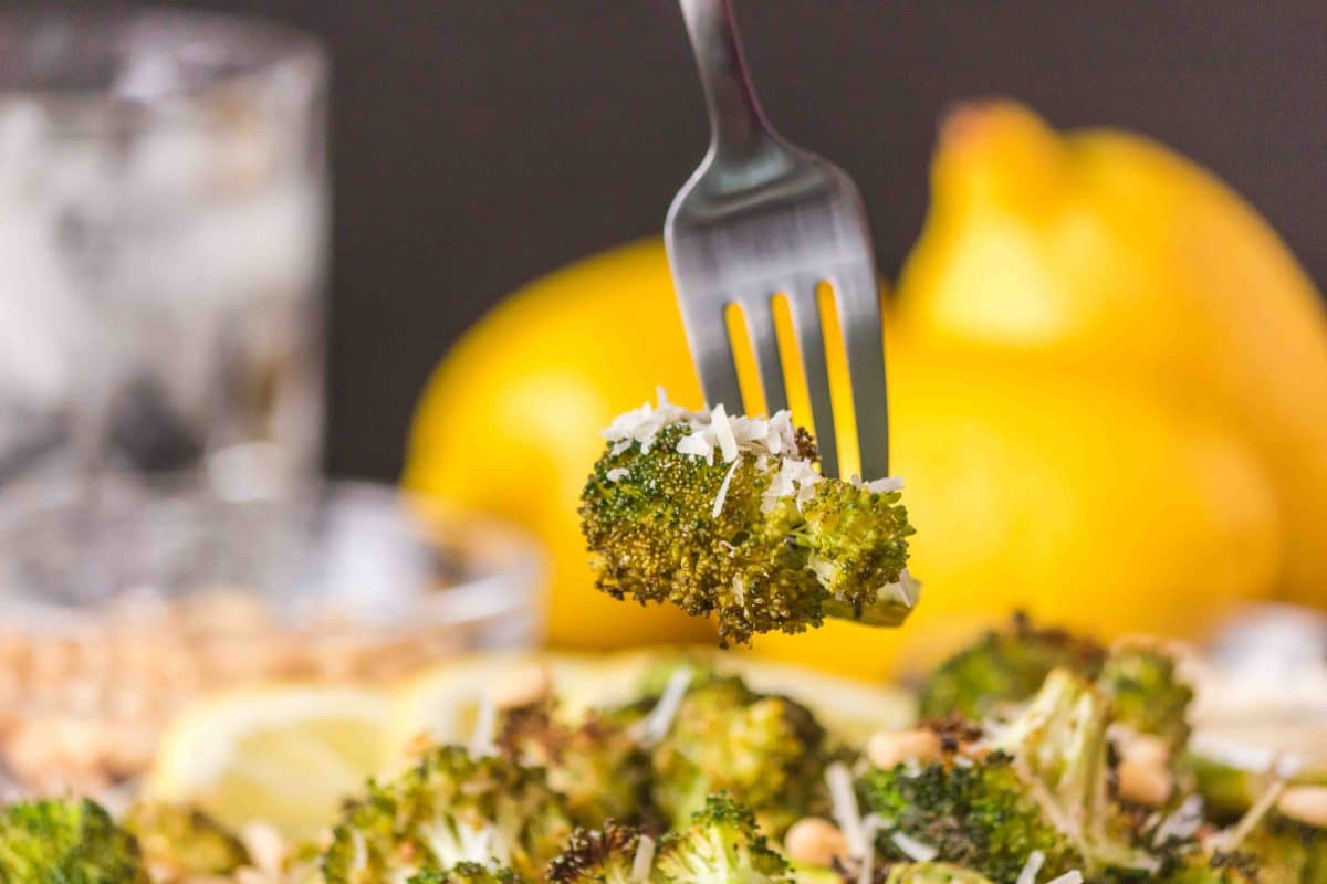 A lemon roasted broccoli floret garnished with parmesan speared with a fork over the serving dish. Hostess At Heart