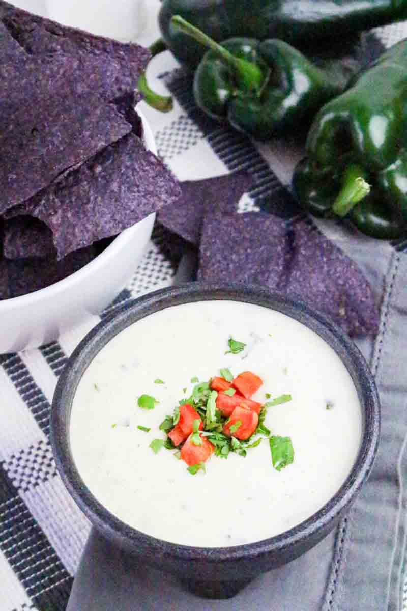 Angled view of a serving bowl filled with queso fundido cheese dip with visible green chili pepper pieces, garnished with chopped tomatoes and cilantro. Hostess At Heart