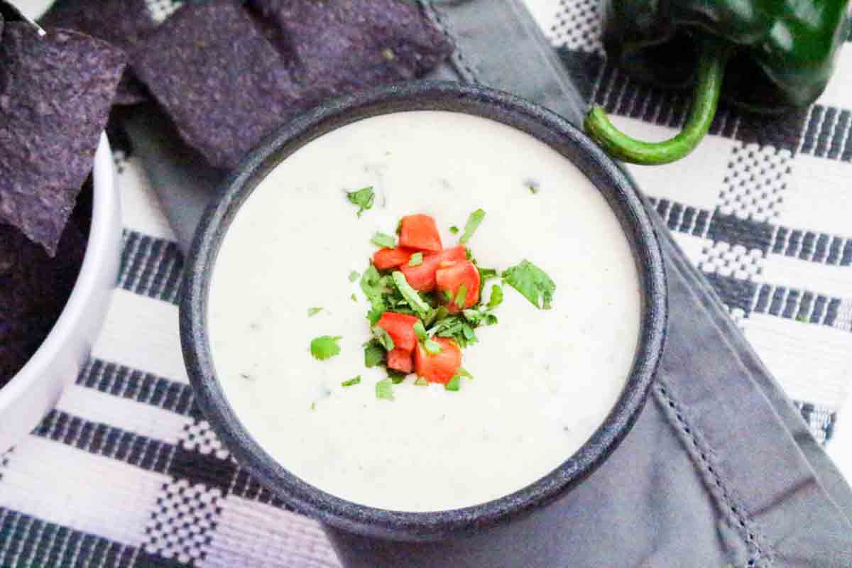 A bowl filled with creamy poblano queso cheese dip garnished with cilantro and tomato. Hostess At Heart