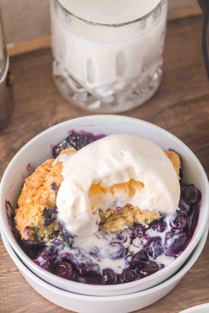 Angled view of a serving of sourdough blueberry cobbler showing the golden topping and jammy blueberry filling underneath with a scoop of vanilla ice cream melting into it. Hostess At Heart