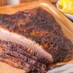 Sideview of a oven baked brisket with a juicy crust with 5 slices laying in front of it on a cutting board. Hostess At Heart