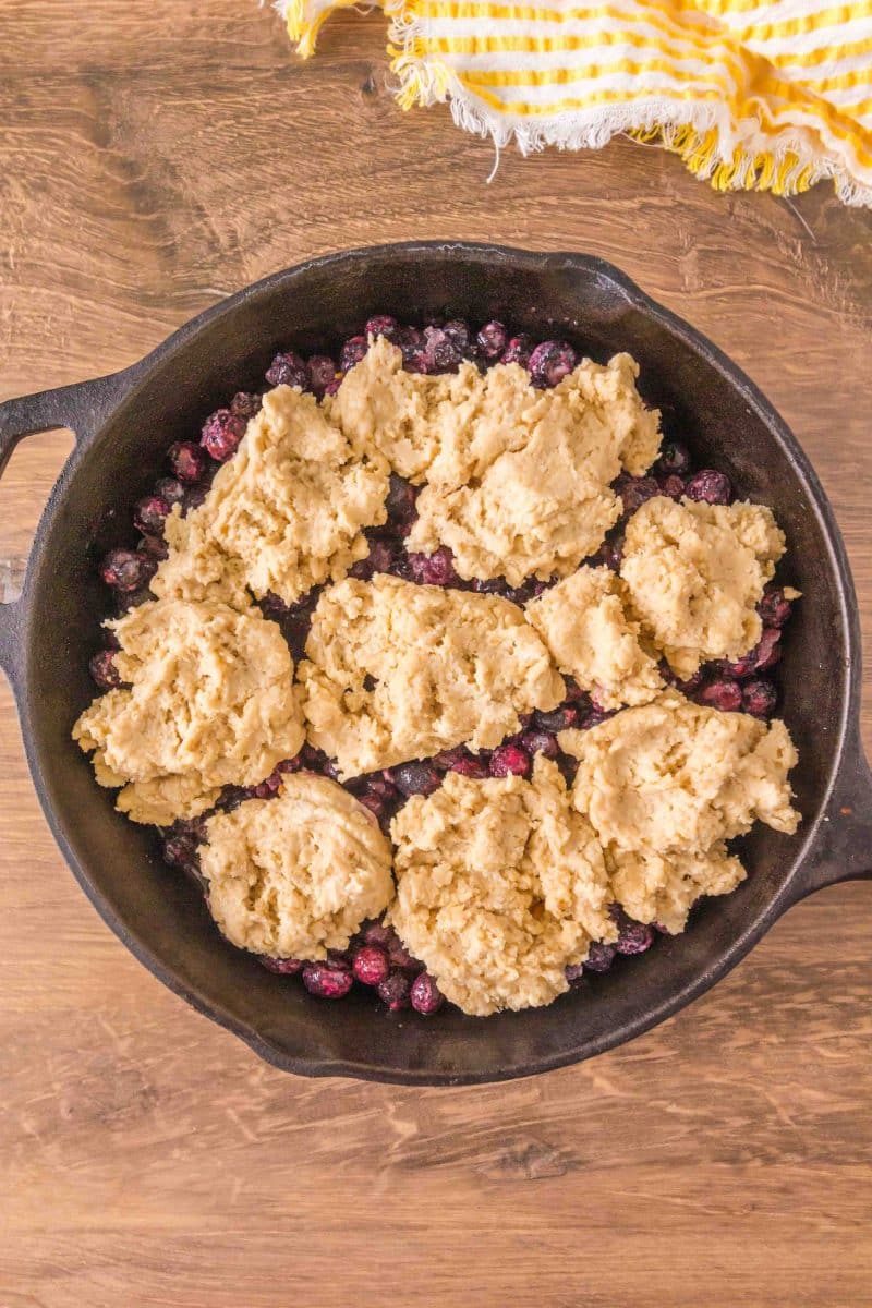 Spoonfulls of sticky sourdough discard dough over blueberry filling in a cast iron skillet ready for baking. Hostess At Heart