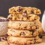 A stack of crumble chocolate chip cookie recipes on a wooden surface. The top cookie is broken in half showing thick soft centers with melted chocolate chips. Hostess At Heart