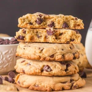 A stack of crumble chocolate chip cookie recipes on a wooden surface. The top cookie is broken in half showing thick soft centers with melted chocolate chips. Hostess At Heart