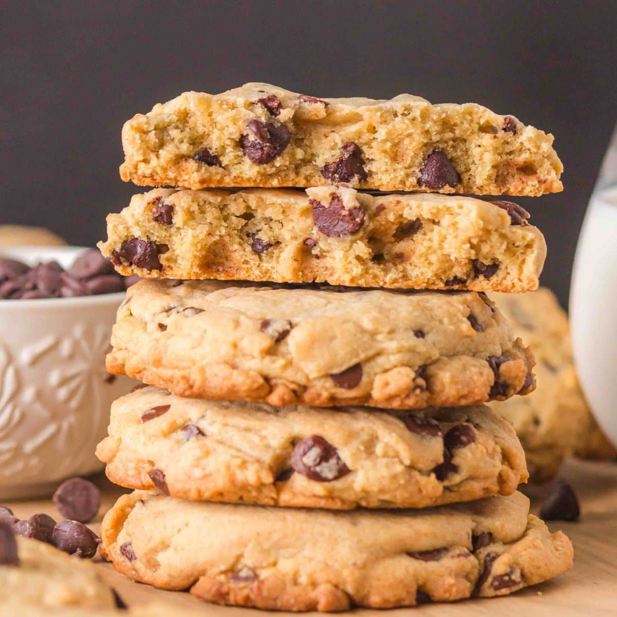 A stack of crumble chocolate chip cookie recipes on a wooden surface. The top cookie is broken in half showing thick soft centers with melted chocolate chips. Hostess At Heart