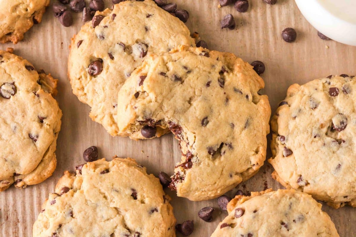 Crumble chocolate chip cookies recipe on a wooden surface. A bite is missing from the center one showing soft melted chocolate in a soft thick center. Hostess At Heart