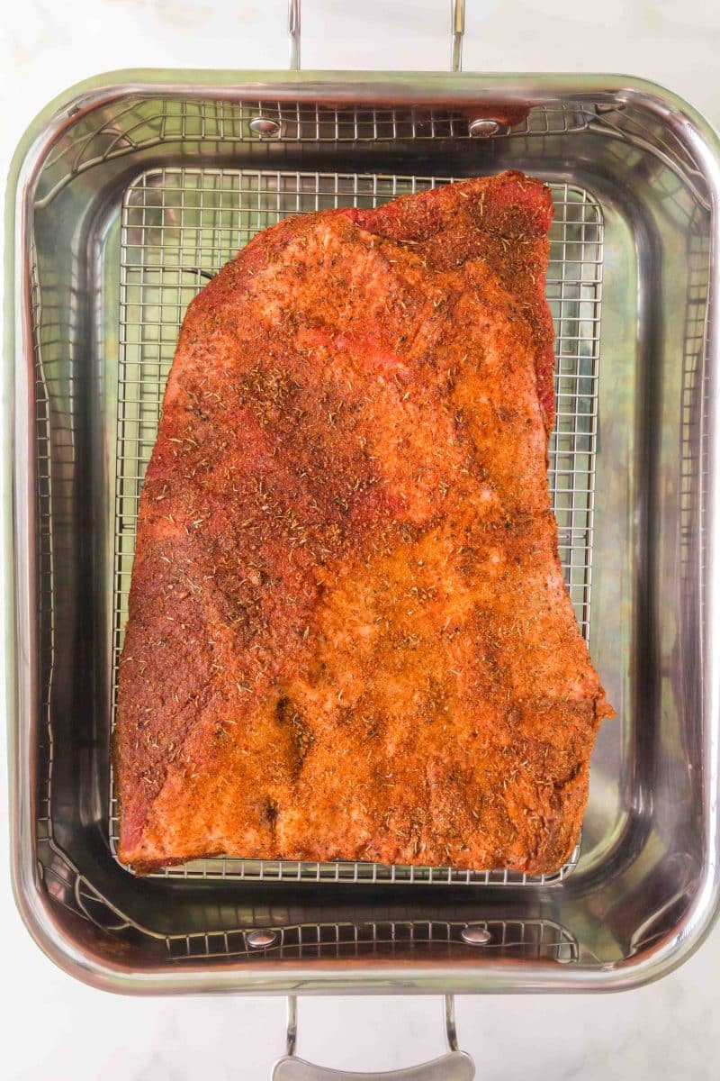 A beef brisket that's been dry rubbed with spices sitting on a baking rack over a roaster ready to be put into the oven. Hostess At Heart