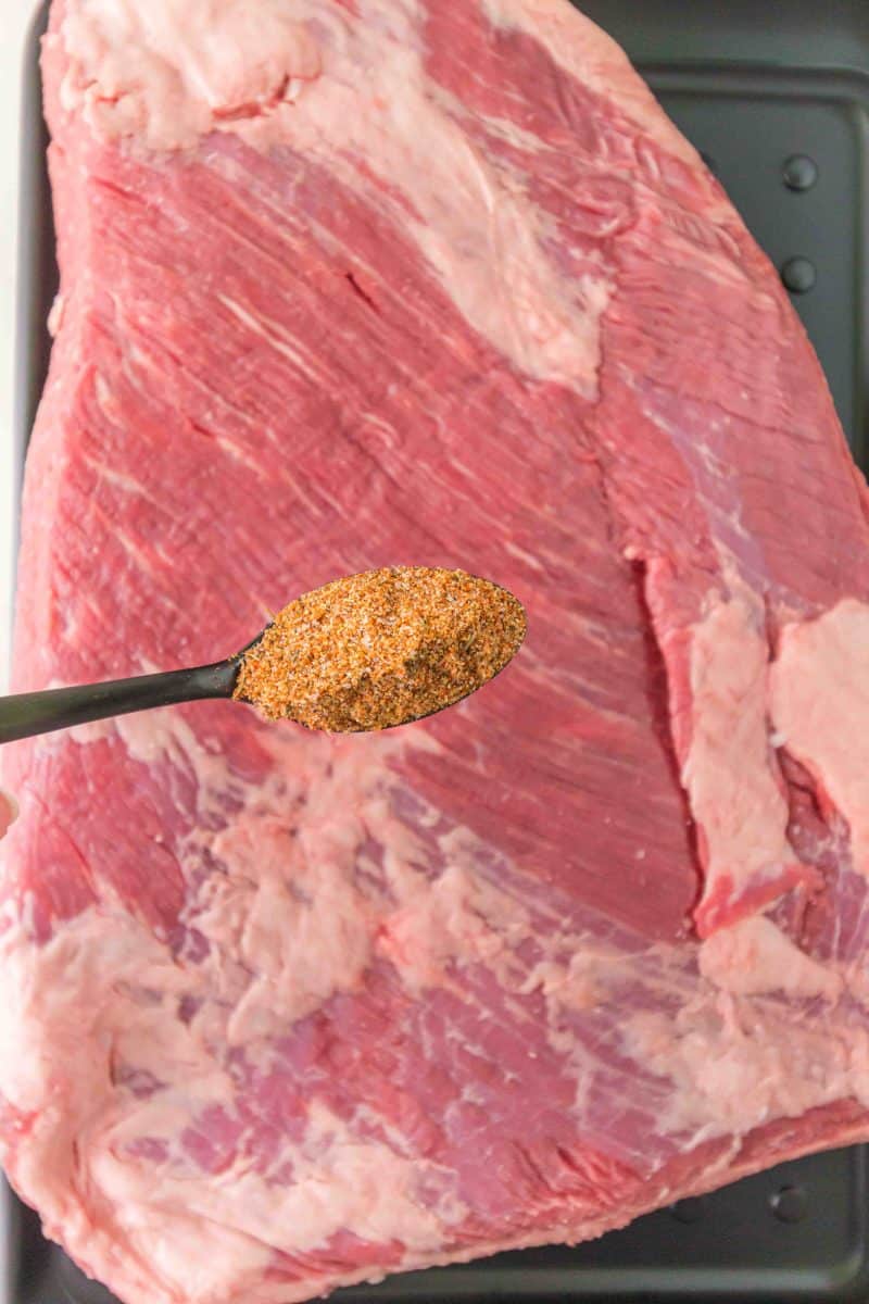 A spoon adding dry rub to a raw beef brisket before baking. Hostess At Heart