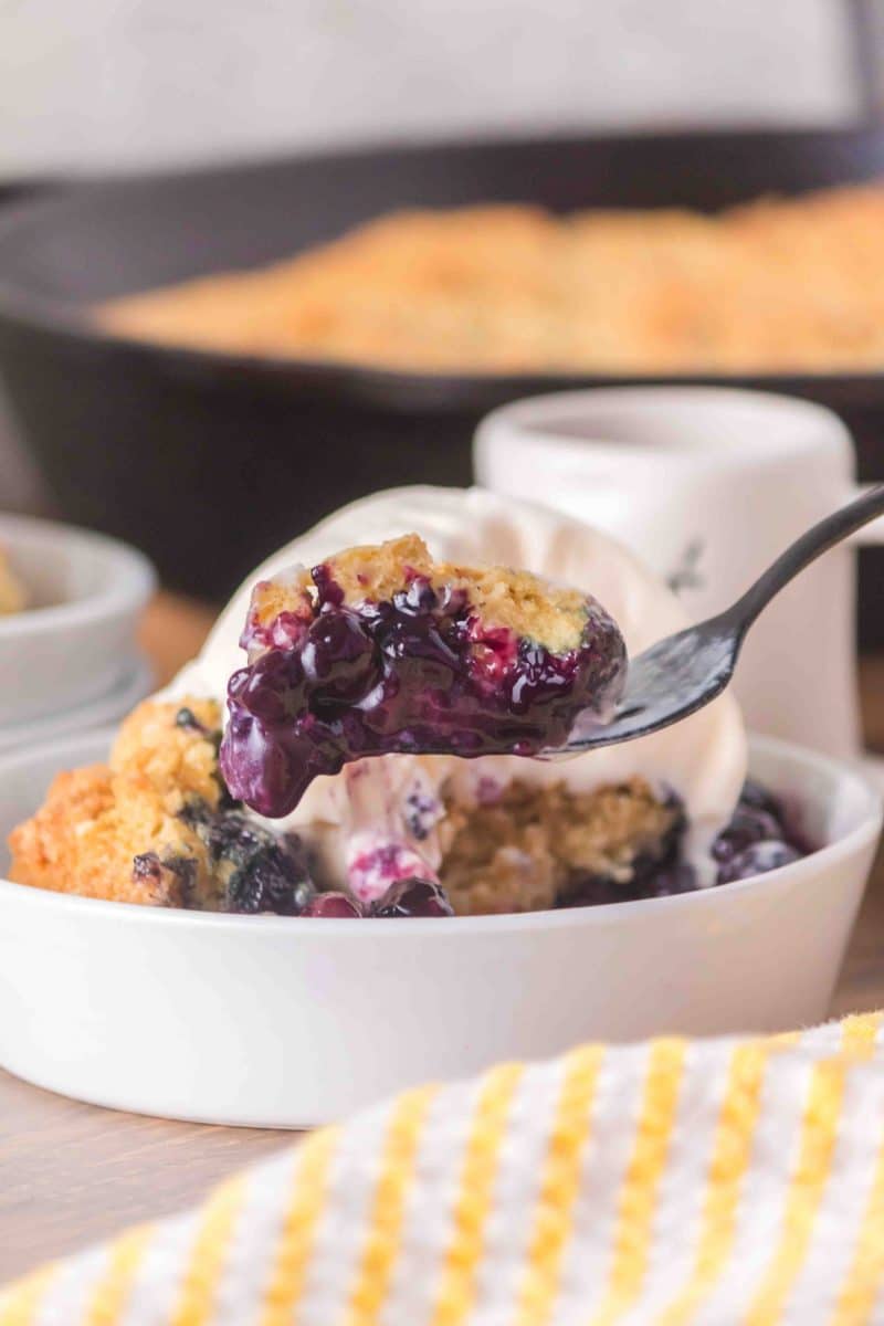 Close-up of a fork filled with sourdough blueberry cobbler showing the golden biscuit topping and a jammy blueberry filling. Hostess At Heart