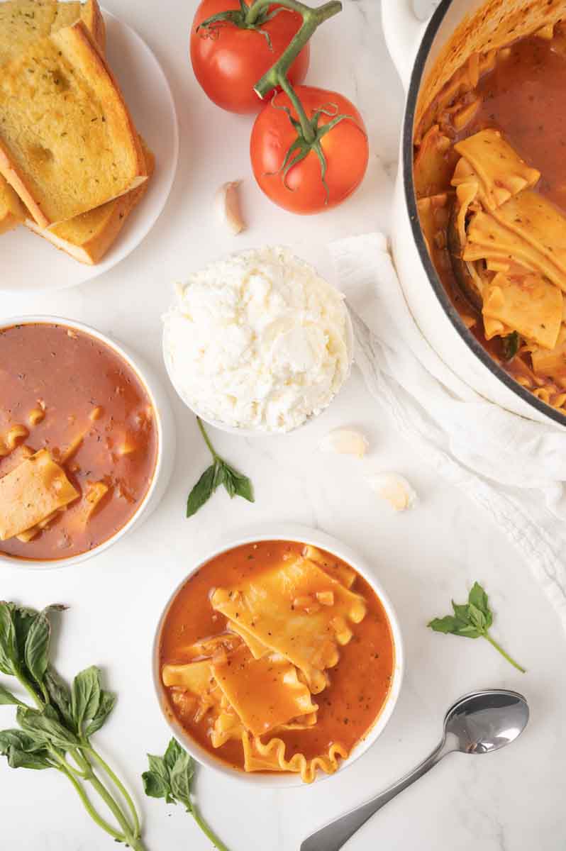 Overhead view of two white bowls of comfort food lasagna soup surrounded by fresh basil, garlic cloves, a bowl of a mixture Ricotta and mozzarella, and a plate of bread, next to the pot of soup. Hostess At Heart