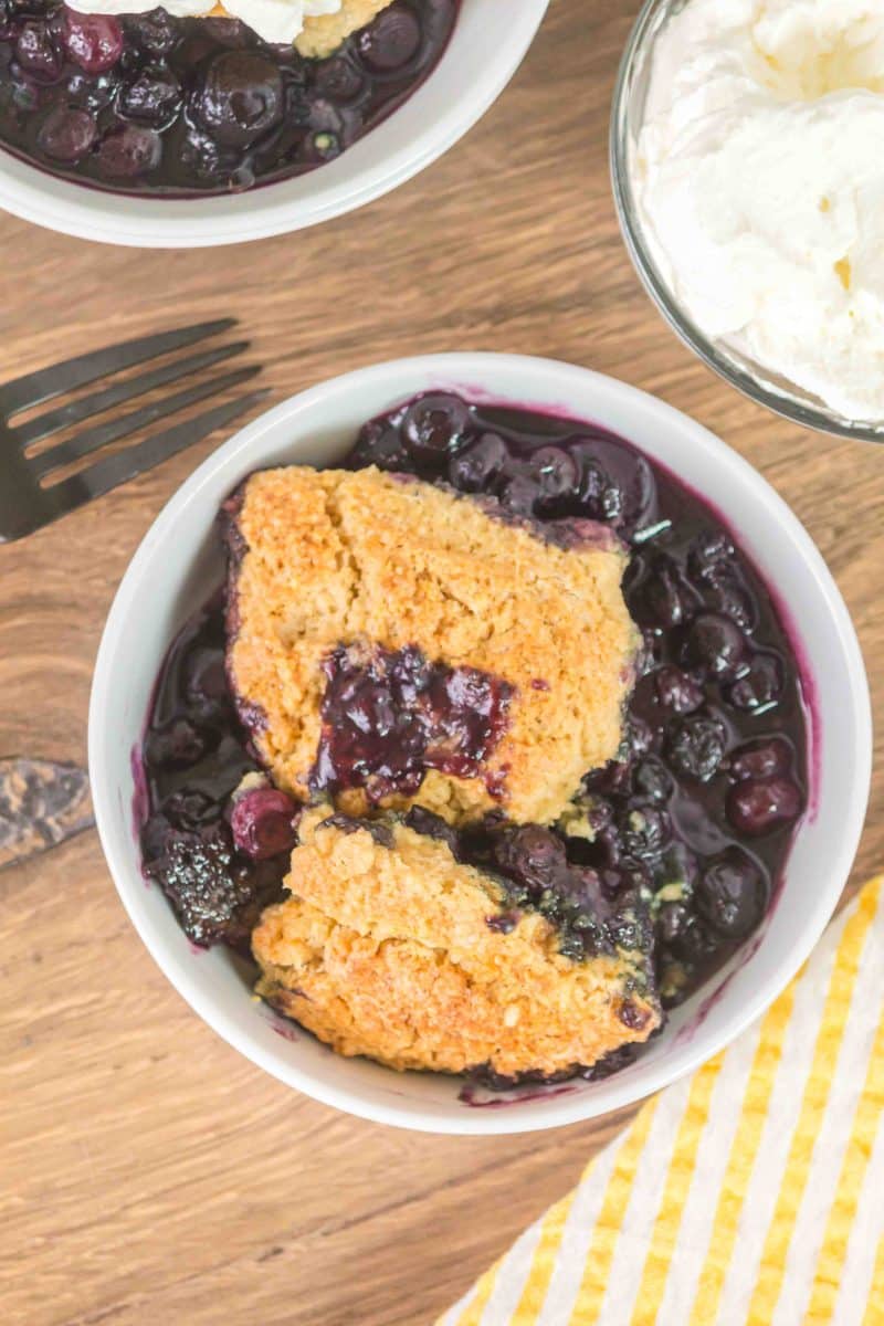 A white bowl filled with baked sourdough discard blueberry cobbler showing a golden baked crust over jammy filling. Hostess At Heart