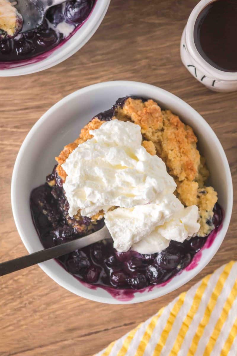 A fork lifting a bite from a white bowl filled with a serving of warm sourdough discard blueberry cobbler topped with a dollop of fresh whipped cream melting into the golden crust and jammy filling. Hostess At Heart