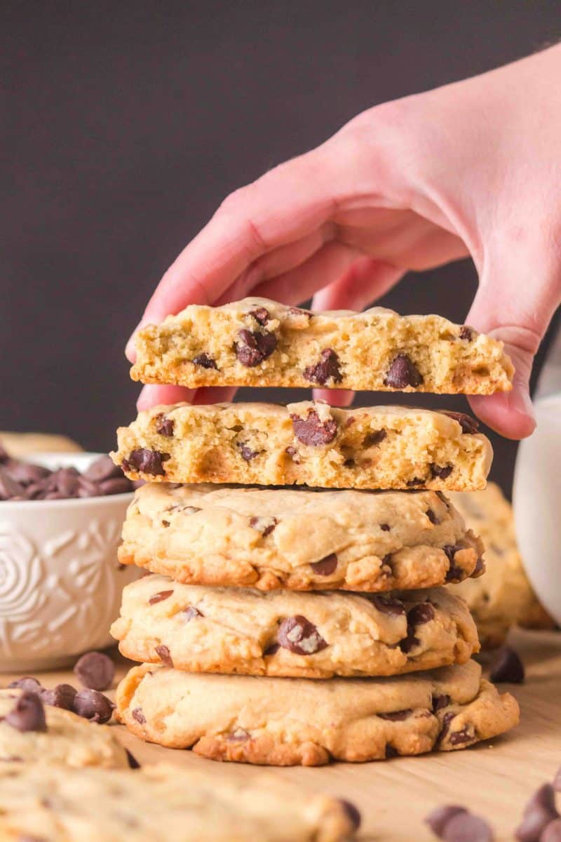 Sideview of a stack of thick crumble chocolate chip cookies. A hand is grabbing the top cookie that's been split in half showing a soft tender center loaded with creamy chocolate chips. Hostess At Heart