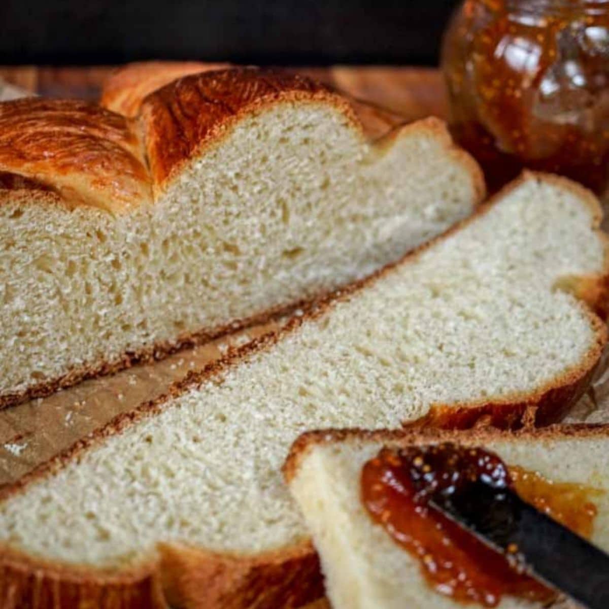 A loaf of Hungarian Egg Twist Bread with the front slices cut from the front. A jar of jam sits in the background and a knife with jam sits on the front slice of bread. Hostess At Heart