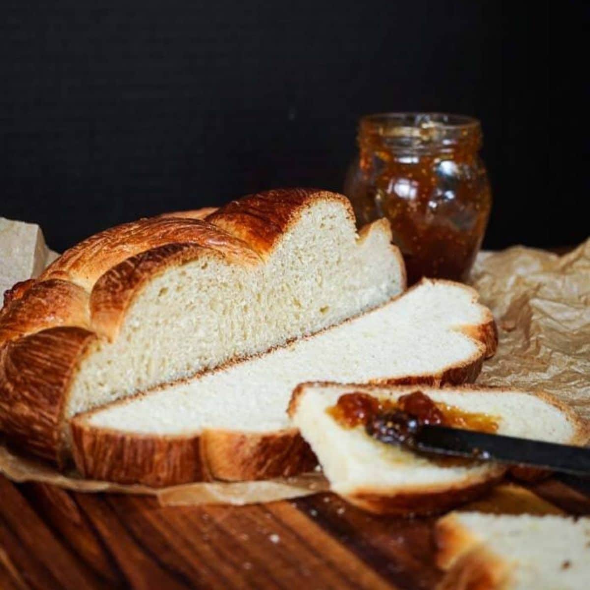 A loaf of Hungarian Egg Twist Bread with the front slices cut from the front. A jar of jam sits in the background and a knife with jam sits on the front slice of bread. Hostess At Heart