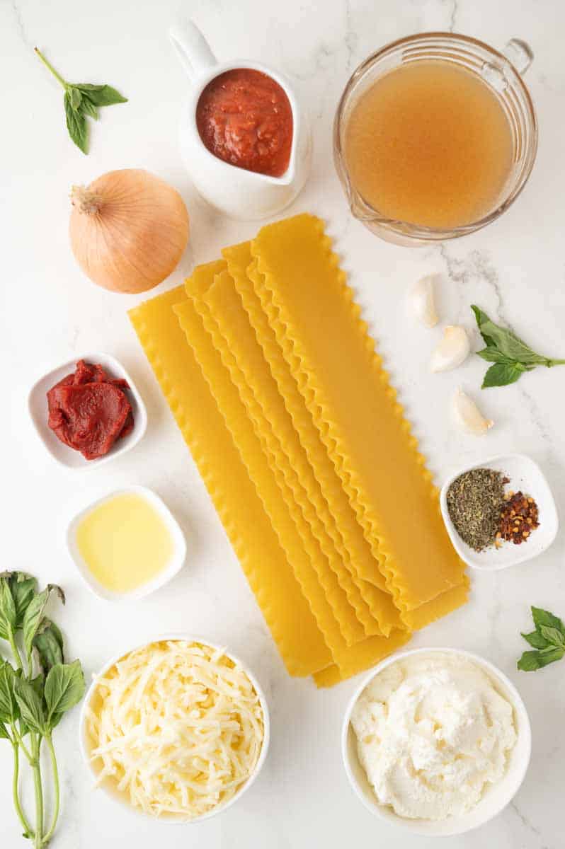 Overhead view of the ingredients needed to make an easy lasagna soup recipe: lasagna noodles, ricotta cheese, mozzarella cheese, Italian seasoning, crushed red pepper flakes, olive oil, tomato paste, onion, vegetable broth, fresh basil, and marinara sauce. Hostess At Heart