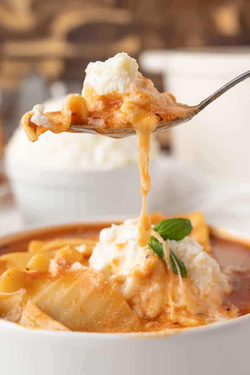 A spoon lifting a cheesy bite of lasagna soup from a white bowl. Hostess At Heart