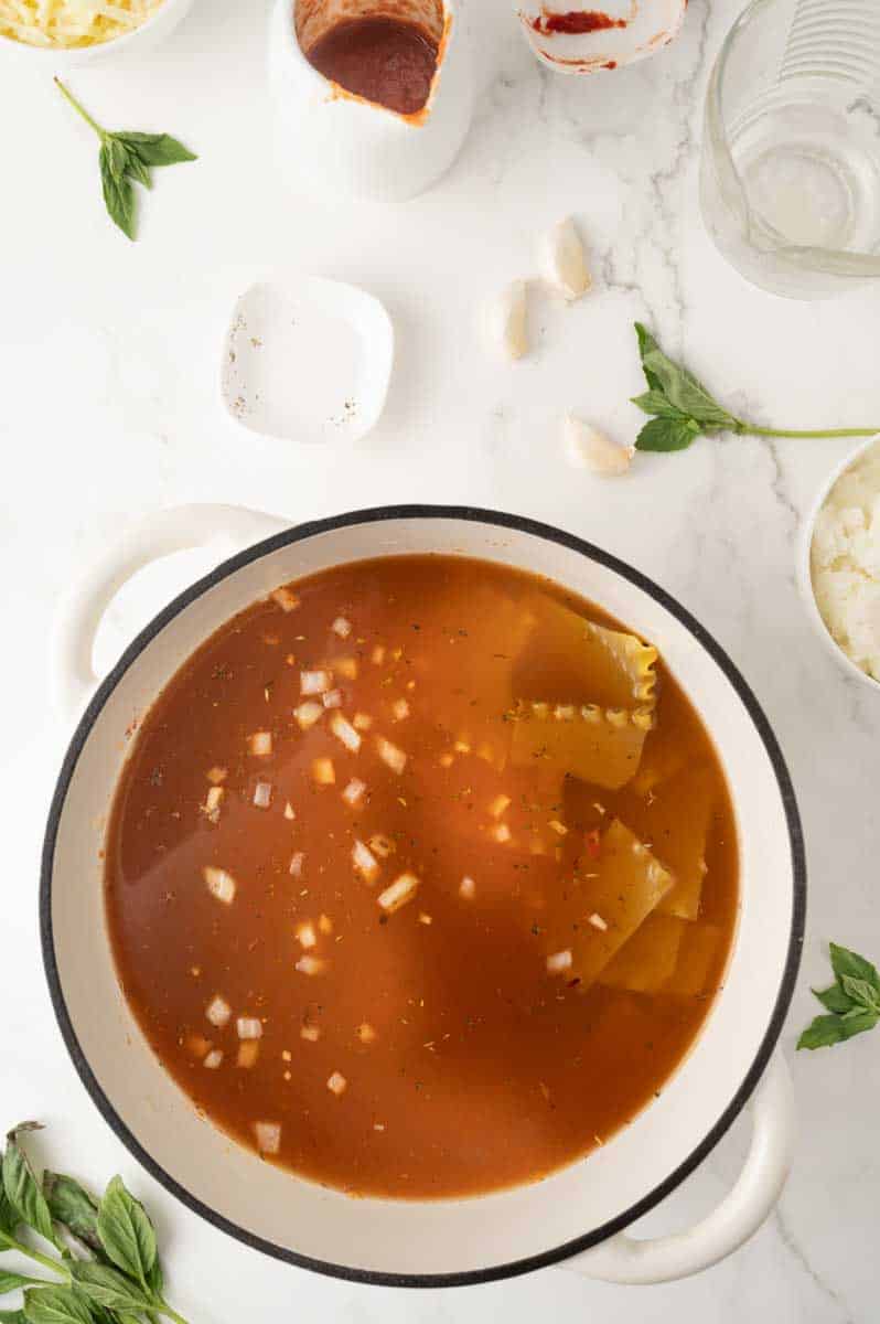 A dutch oven filled with a mixture of tomato vegetable broth, minced garlic, and chopped onion to make a vegetarian lasagna soup recipe. Hostess At Heart