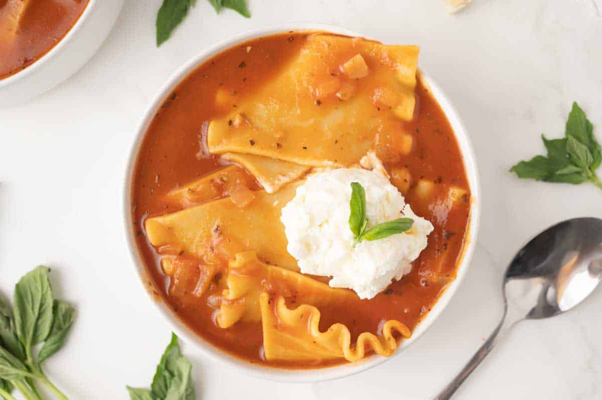 Top down view of a bowl of easy lasagna soup topped with ricotta mozzarella cheese mixture and fresh basil leaves. Hostess At Heart