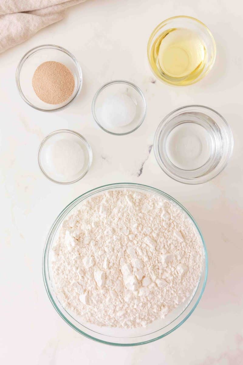 Ingredients for no knead dough recipe including flour, yeast, olive oil, sugar, salt, and warm water in bowls and arranged on a white surface. Hostess At Heart
