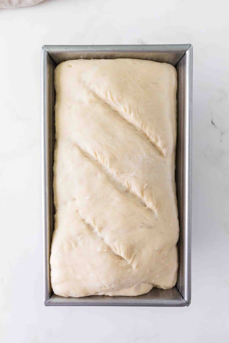 Easy no knead dough rising in a bread loaf pan, scored and ready for baking. Hostess At Heart