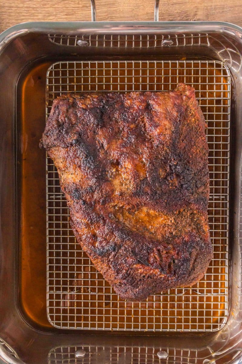 Top down view of a golden brown beef brisket fresh from the oven sitting on a baking rack above the juice in the pan. Hostess At Heart