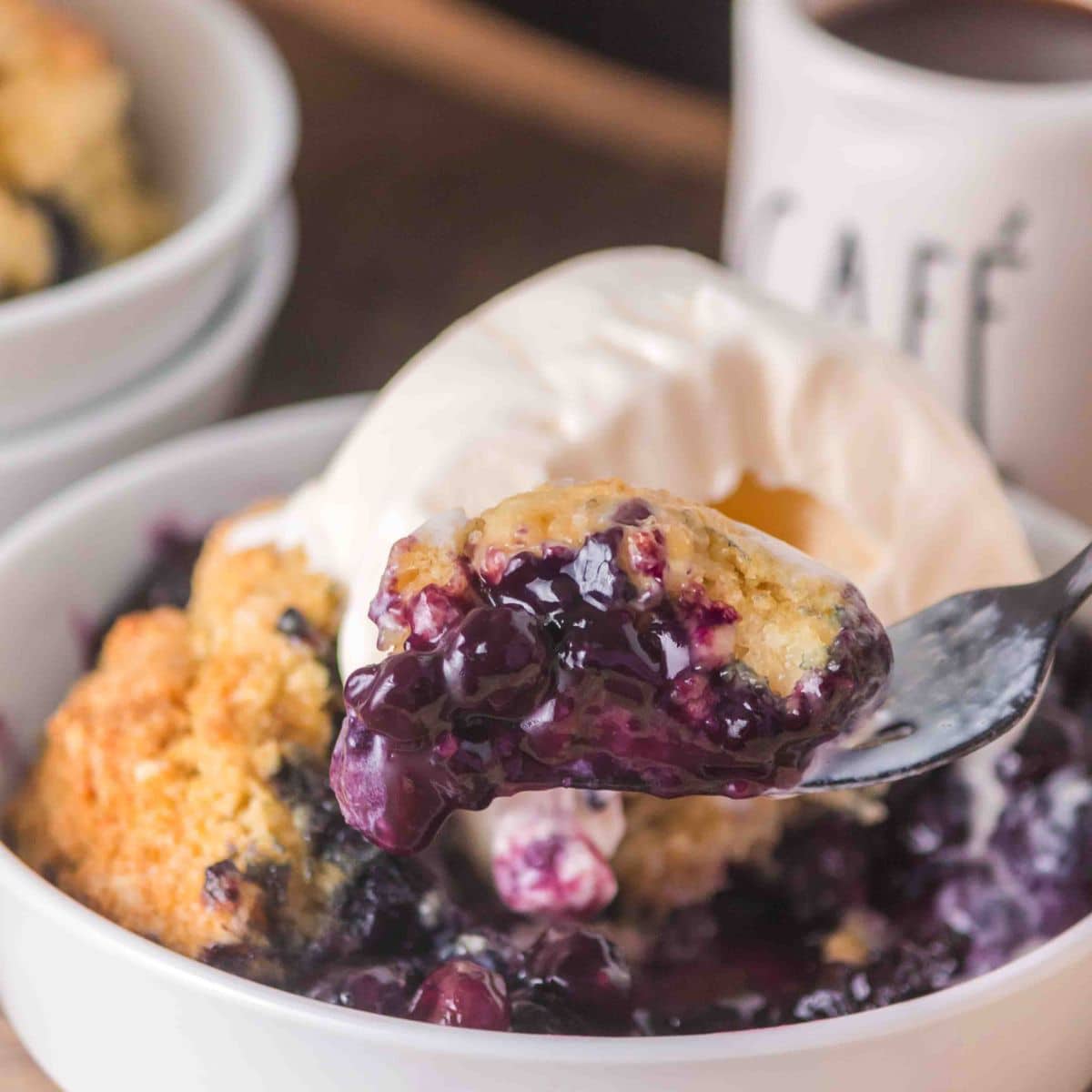 A serving of warm sourdough discard blueberry cobbler in a white bowl topped with a scoop of vanilla ice cream, melting into the golden crust with a fork lifting out a bite. Hostess At Heart