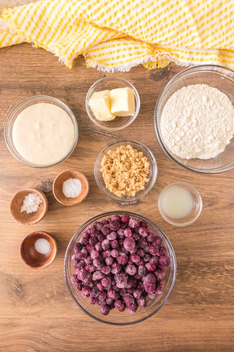 Ingredients for sourdough discard blueberry cobbler including frozen blueberries, sourdough discard, lemon juice, salt, butter, flour, baking soda, baking powder, and brown sugar arranged on a wooden surface. Hostess At Heart