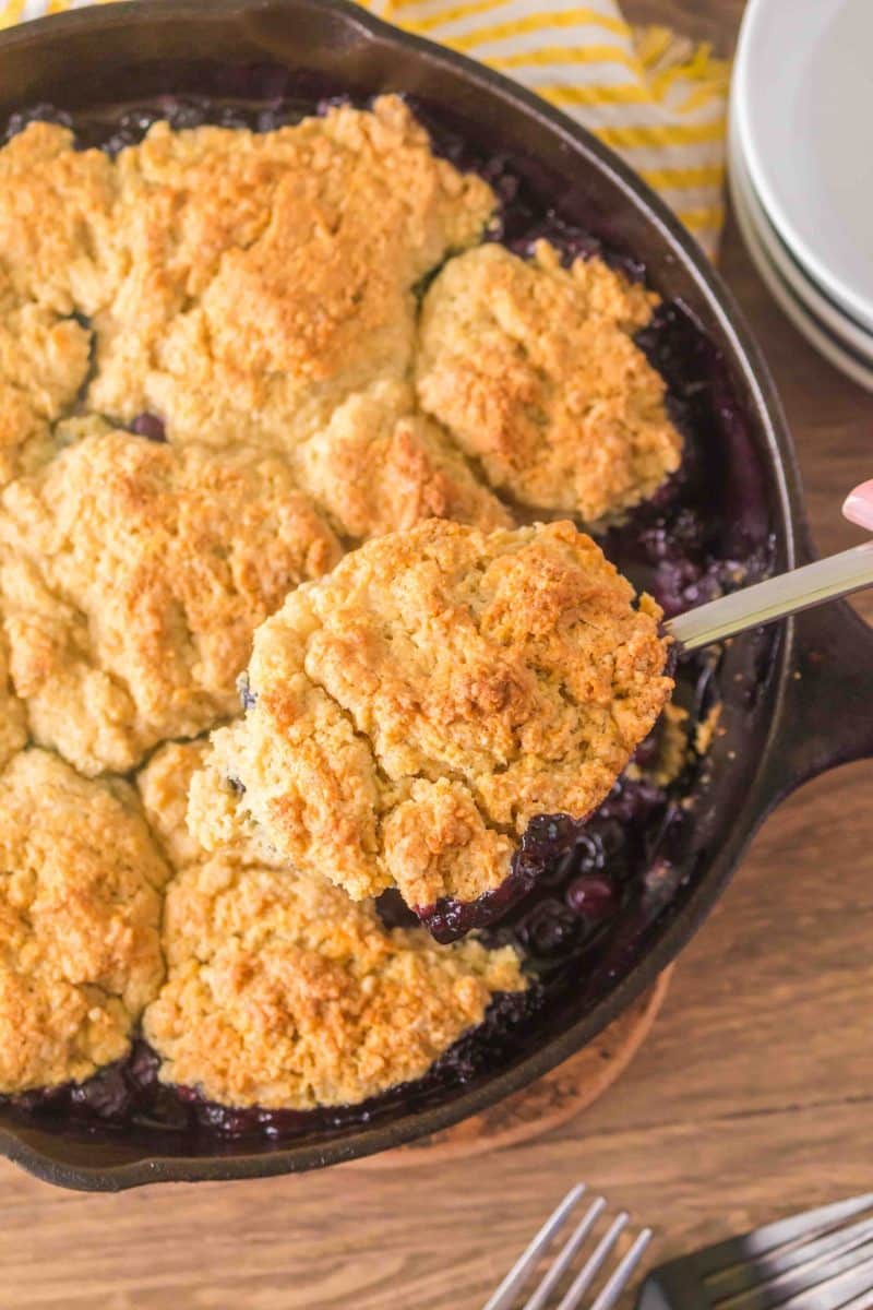 Golden Sourdough discard blueberry cobbler in a cast iron skillet with a serving lefted out of a serving spatula. Hostess At Heart