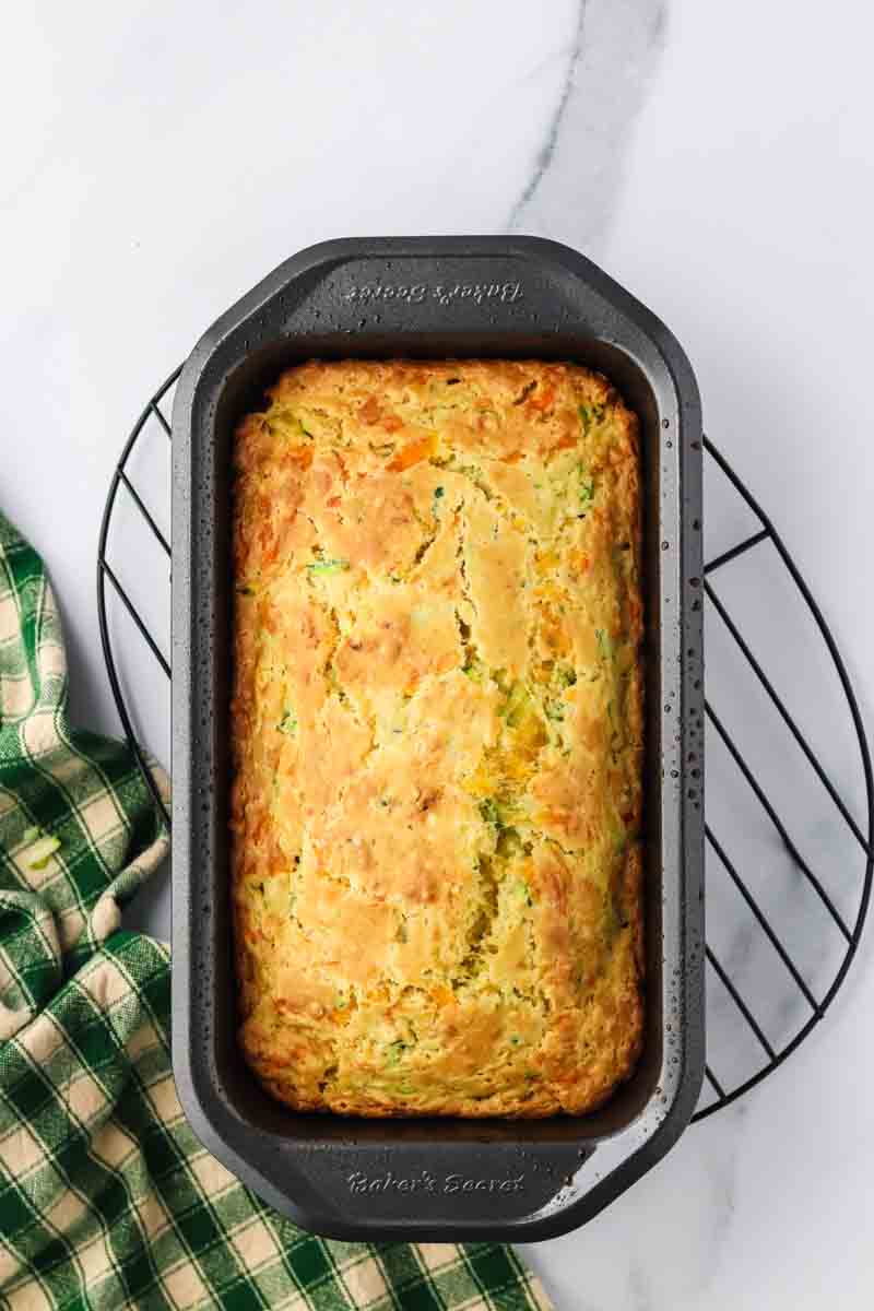 Baked Zucchini Cheese Bread fresh from the oven waiting to be topped with shredded cheddar cheese in a loaf pan. Hostess At Heart