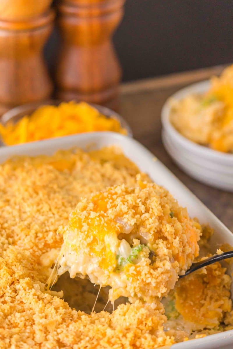 A spoonful of broccoli rice casserole being lifted from a white baking dish showing melted cheese and crispy cracker topping. Hostess At Heart