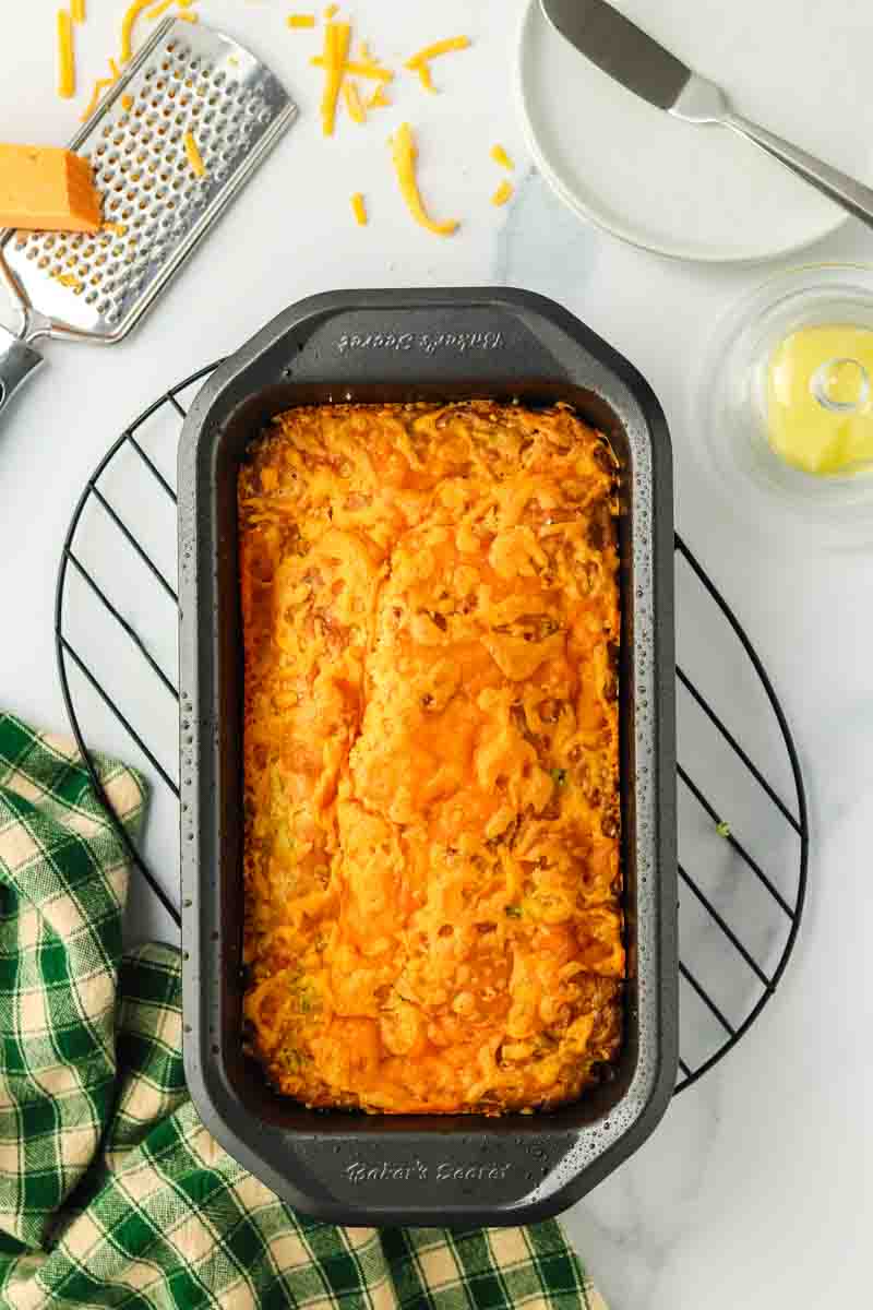 Baked zucchini cheese bread with golden cheddar topping in a loaf pan cooling on a wire rack. Hostess At Heart