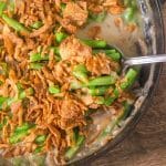 A spoon lifting a serving of Green Bean Casserole with fresh green beans showing fresh cut green beans in a thick mushroom sauce topped with crispy fried onions. Hostess At Heart
