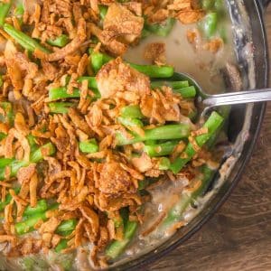 A spoon lifting a serving of Green Bean Casserole with fresh green beans showing fresh cut green beans in a thick mushroom sauce topped with crispy fried onions. Hostess At Heart