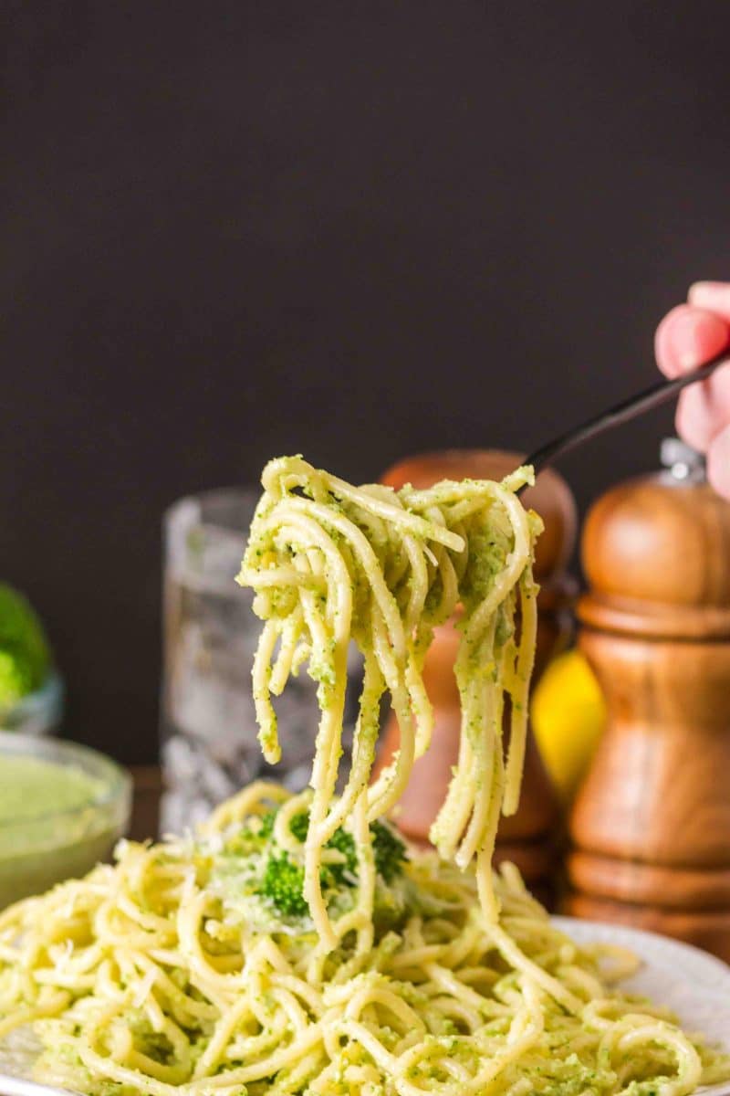 A fork lifting a serving of broccoli pesto spaghetti from a white serving bowl. Hostess At Heart