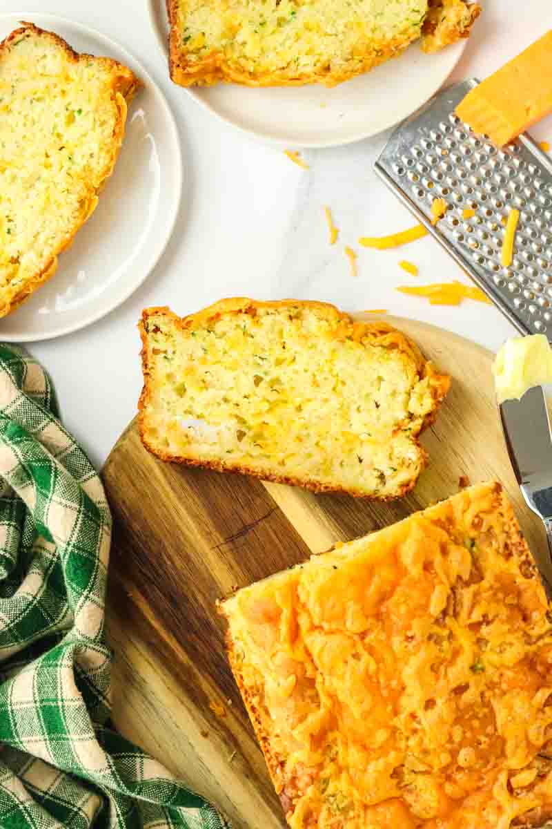 Overhead view of a partially sliced loaf of zucchini cheese bread sitting on a wooden cutting board with a slice laying in the front showing a soft and tender crumb dotted with shredded zucchini. Hostess At Heart