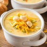 Overhead shot of creamy broccoli cheddar soup in a white bowl topped with shredded cheddar and crackers. Hostess At Heart