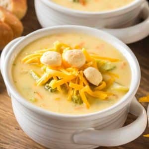 Overhead shot of creamy broccoli cheddar soup in a white bowl topped with shredded cheddar and crackers. Hostess At Heart