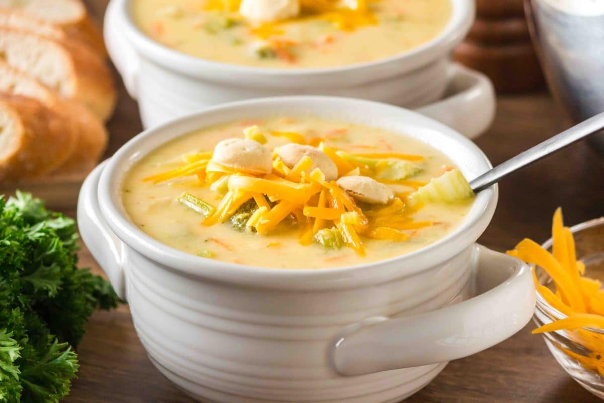 Overhead shot of creamy broccoli cheddar soup in a white bowl topped with shredded cheddar and crackers. Hostess At Heart