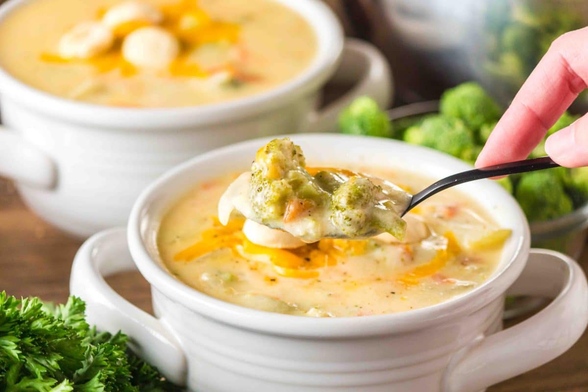 A side shot of a spoon lifting a bite of rich and creamy broccoli cheddar soup in a white bowl. Hostess At Heart