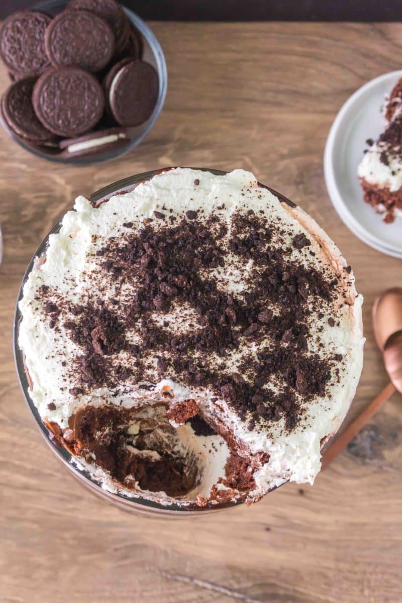Overhead view of a chocolate pudding trifle dessert with a scoop missing showing all of the layers of chocolate cake, pudding, whipped cream and oreo cookie crumbs. Hostess At Heart
