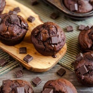 A chocolate pumpkin muffin topped with melted chocolate chunks surrounded by more chocolate pumpkin muffins. Hostess At Heart