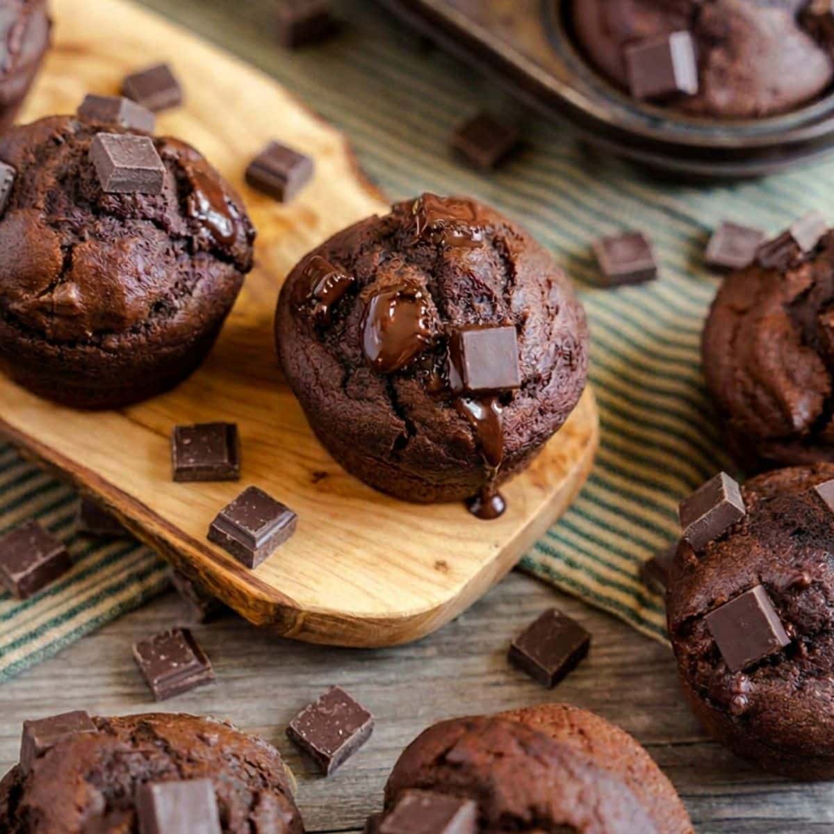 A chocolate pumpkin muffin topped with melted chocolate chunks surrounded by more chocolate pumpkin muffins. Hostess At Heart