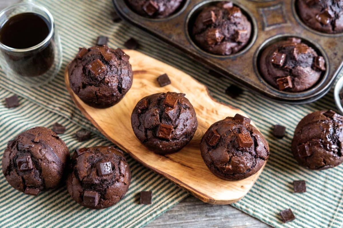 Three-quarter view of baked chocolate pumpkin muffins on a a cutting board with the tin of baked muffins in the background. Hostess At Heart